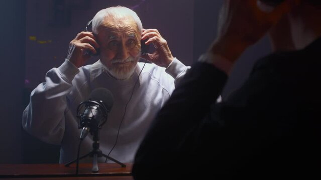 The old man creates a podcast in a professional studio. Senior male does online streaming interviews. Elderly man blogger speaking in microphone or make vlog. Recording audio content for social media