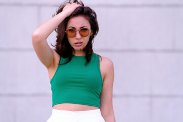 Fototapeta na wymiar Beautiful girl in a green top, white shorts and sunglasses posing. Aggressively, displeased looks at the camera. Against the background of a gray wall