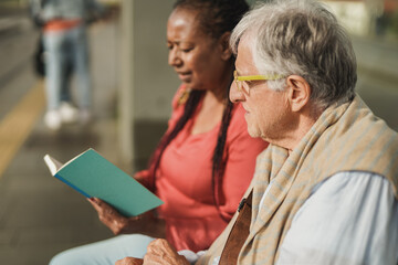 Senior man and african mature woman sitting together at tram station - Elderly nursing care concept