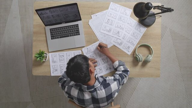 Top View Of Asian Male Artist Drawing Storyboard For The Film On The Table With A Laptop And Headphones In The Studio
