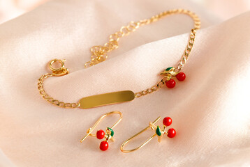 Gold bracelet with a name plate and earrings in the shape of a cherry (red and green enamel) on a pink background. Romantic jewelry. Advertising still life product.