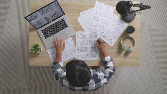 Top View Of Asian Male Artist Looking At Sketching Concept On A Laptop Then Drawing Storyboard On The Table With Headphones In The Studio
