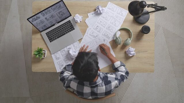 Top View Of Unsatisfied Asian Male Artist Crumples Paper While Drawing Storyboard For The Film On The Table With A Laptop And Headphones In The Studio
