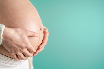 Pregnant woman gently holding her naked round belly with her hands in last month of pregnancy. Third trimester - week 36. Close up. Side view. Turquoise background. Bright shot.