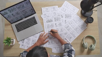 Top View Zoom Out Shot Of Asian Male Artist Drawing Storyboard For The Film On The Table With A Laptop And Headphones In The Studio
