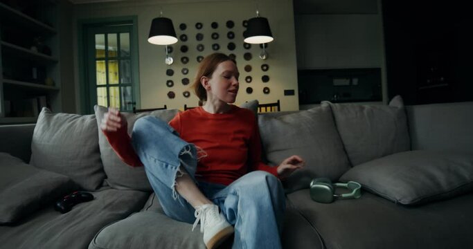 A young woman in casual clothes sits on the sofa, puts on headphones and starts gambling in a video game using a joystick