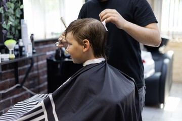 Child boy sitting in a chair in a barbershop carefully watching the work of a hairdresser in the...