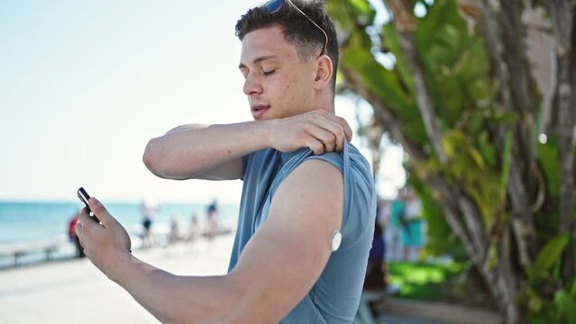Young hispanic man tourist measuring glucose using smartphone and freestyle at seaside