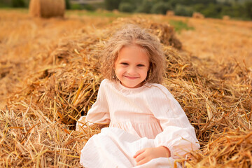 Naklejka na ściany i meble a blonde girl with long hair in a pink linen dress is sitting on a haystack in a mown field