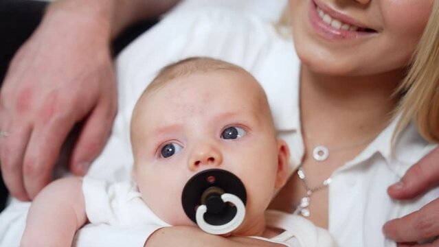 Grey-eyed Caucasian newborn with a black pacifier. Mom waves her son and kisses him on the head. Close up.