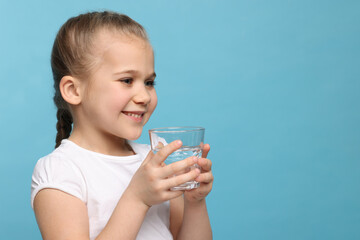 Happy little girl holding glass of fresh water on light blue background, space for text