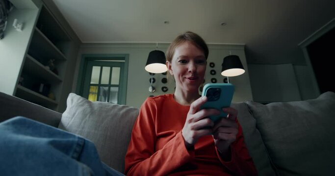 A young red-haired woman in casual clothes laughs while typing on a mobile phone while sitting on a sofa in a lotus pose. Home interior