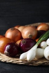 Wicker mat with different kinds of onions on black wooden table, closeup