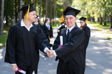 A group of graduates in robes outdoors. An elderly man and a young guy congratulate each other on receiving a diploma.