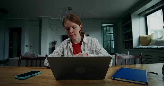 A young red-haired woman in casual clothes puts on headphones and starts a video chat, sitting in front of an open laptop. She smiles and waves hello