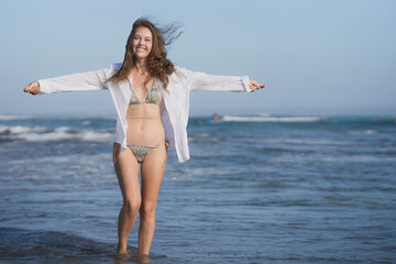 A slender European woman with brown hair in a white shirt and a blue bikini is having fun on the ocean coast on a bright sunny day. Rest on the tropical beaches of Asia. Full length photo.