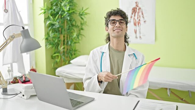 Young hispanic man doctor using laptop holding rainbow flag at clinic
