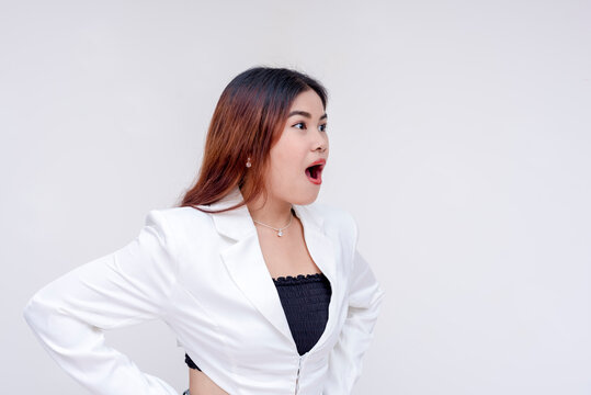 A surprised young woman with raised eyebrows and mouth wide open facing sideways to the right with hands on her hips. Isolated on a white background.