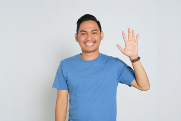 Happy young Asian man in blue t-shirt showing hi gesture with waving hand and smiling isolated on...