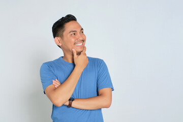 Smiling young Asian man in blue t-shirt looking at copy space, touching his chin, interested in good sale offer isolated on white background