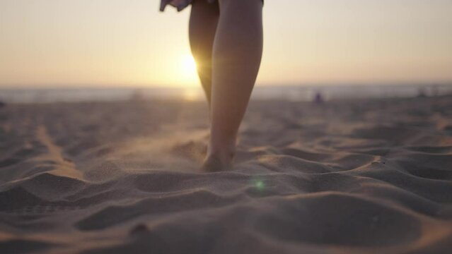 Woman walks barefoot on sandy beach toward sunset, slomo forward tracking
