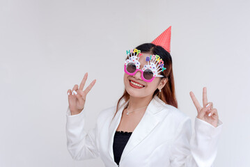 A cheerful young woman doing peace sign with both hands while wearing a happy birthday sunglasses and a pink party hat. Isolated on a white background.