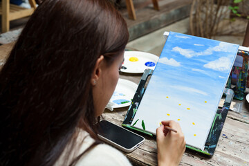 Asian girls spend their free time on vacation. Use your hand to paint the paint brush on the fabric stretched on the wooden frame. Practicing drawing, making art work painting of flowers and sky