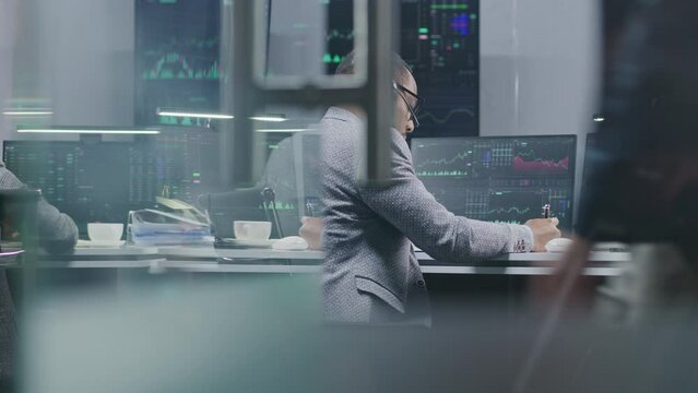 African American financial analyst monitors real-time stocks on computers, writes data with pen, works in bank office. Exchange market charts displayed on big digital screens. Investment or analytics.