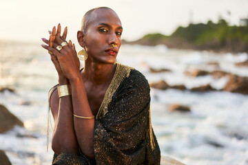 Epatage lgbtq black man poses on scenic ocean beach looks at camera demonstrates jewellery . Androgynous ethnic fashion model in posh dress looks at camera, sits on stone in - obrazy, fototapety, plakaty