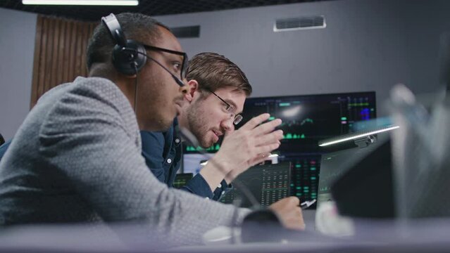 Mixed race stock traders talk and monitor real-time stocks data on computer. Colleagues work in broker agency office. Big digital screen displayed exchange market charts. Investment and analytics.