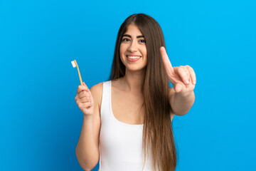 Young caucasian woman brushing teeth isolated on blue background showing and lifting a finger