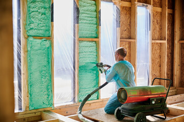 Male builder insulating wooden frame house. Man worker spraying polyurethane foam inside of future...