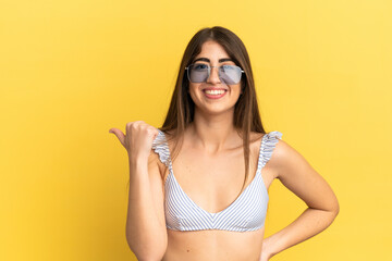 Young caucasian woman in swimsuit in summer holidays isolated on yellow background pointing to the side to present a product