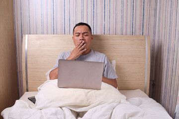 Fototapeta premium Asian Guy yawning while using laptop in comfortable bed in bedroom at home.