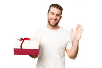 Young handsome blonde man holding a gift over isolated chroma key background saluting with hand with happy expression