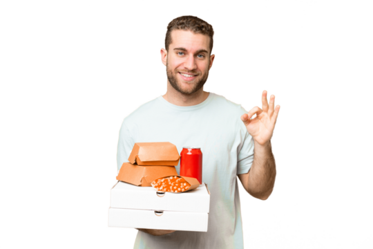 Young handsome blonde man holding pizzas and burgers over isolated background showing ok sign with fingers