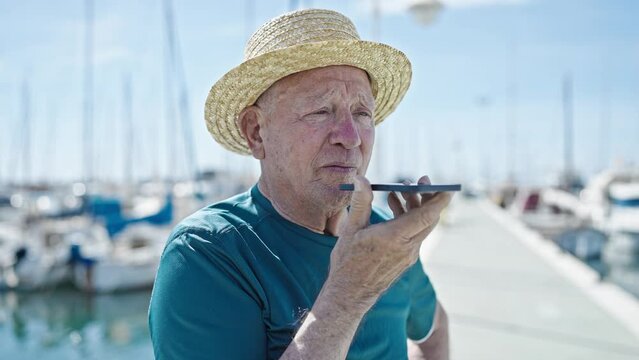 Senior grey-haired man tourist wearing summer hat sending voice message by smartphone at port
