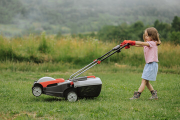 Fototapeta premium A child in boots in the form of a game mows grass with a lawnmower in the yard against the background of mountains and fog, the concept of garden tools