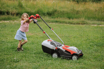 A child in boots in the form of a game mows grass with a lawnmower in the yard against the background of mountains and fog, the concept of garden tools