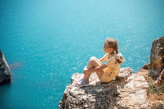 Happy girl perched atop a high rock above the sea, wearing a yellow jumpsuit and braided hair, signifying the concept of summer vacation at the beach.