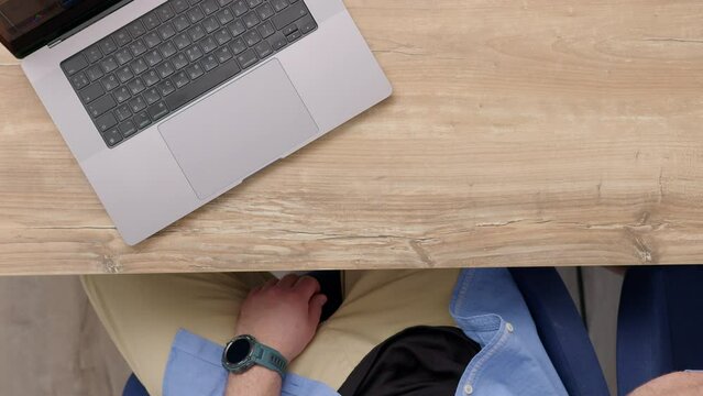 unrecognizable worker with blue shirt sits at his desk, drinks coffee and watch video on laptop. Office worker or freelancer works behind computers, top down view. Work from office or from home.
