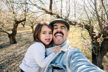 A selfie of a father and his young daughter making funny gestures in a field of almond blossoms