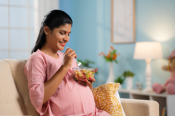 Happy indian pregnant woman eating fruit salad while sitting on sofa at home - concept of Pregnancy...
