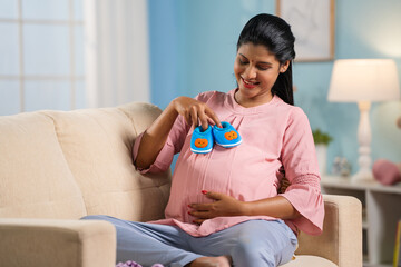 Happy indian pregnant woman playing with baby shoes or booties on tummy at home - concept of...