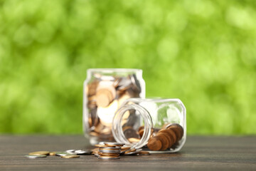 Stack of coins and jars on wooden table against blurred green nature background. Savings concept