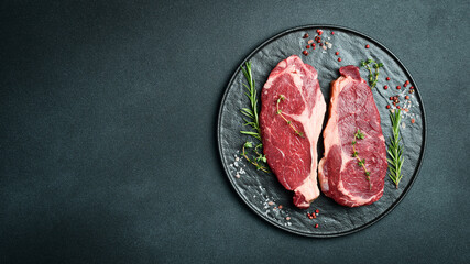 Two raw rib eye beef steaks with spices on a plate. Ready to cook. On a dark stone background. Top...