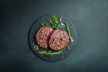 Raw burger patty with spices and herbs on a slate plate. On a dark stone background. Top view.