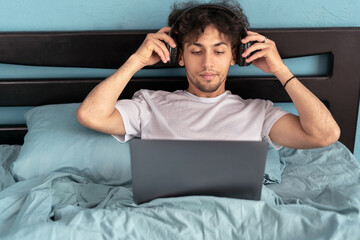 Man using laptop wearing headphones watching movie online sitting in bed in bedroom at home. Weekend leisure and freelance lifestyle
