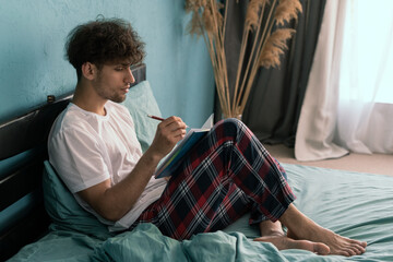 Handsome young man taking notes in notebook while lying on bed writing down ideas and doing planning
