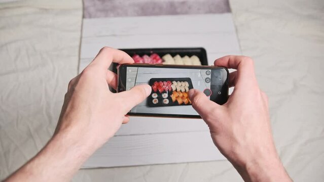 Blogger taking a photo of sushi rolls by a smartphone camera for posting on social networks. Male hands filming Japanese food in a delivery plastic box on a mobile phone. Staged shot for social media.